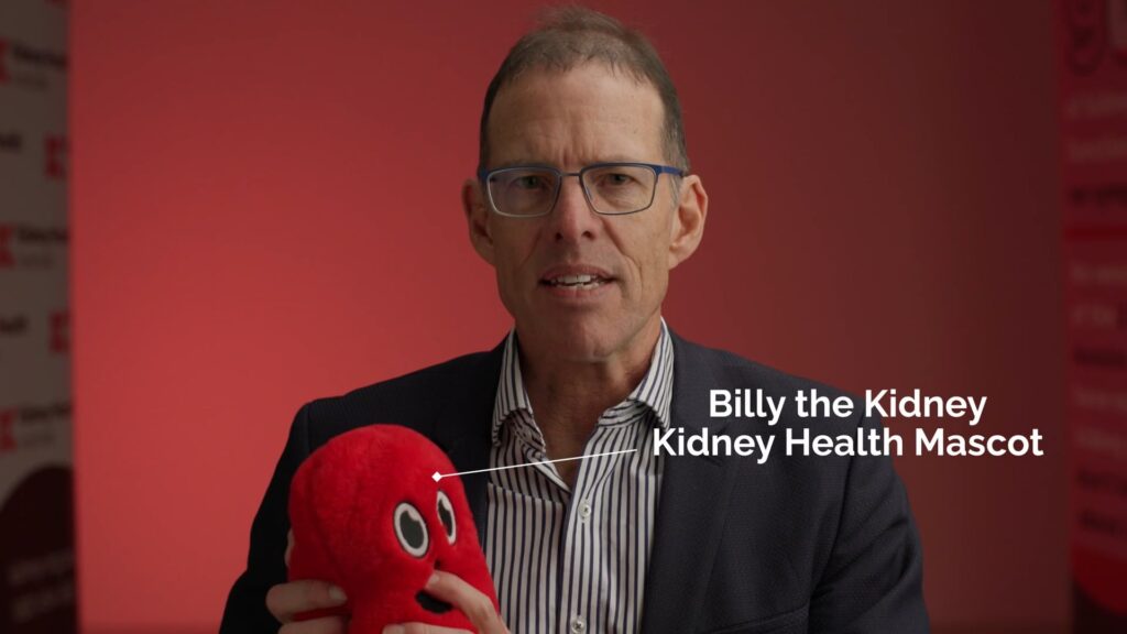 Man in glasses speaking to camera against a red backdrop, holding a red plush toy kidney labelled “Billy the Kidney, Kidney Health Mascot.”