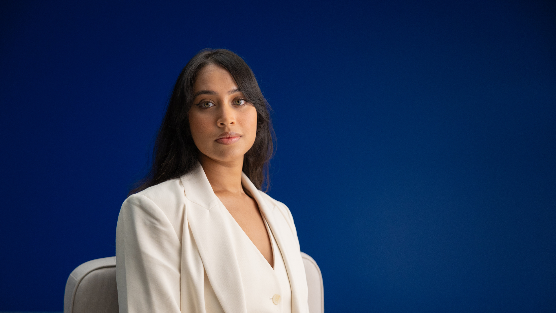 A professional woman in a white blazer sits in front of a bold, blue background during a video shoot for the Legal Services Commission of South Australia recruitment campaign.