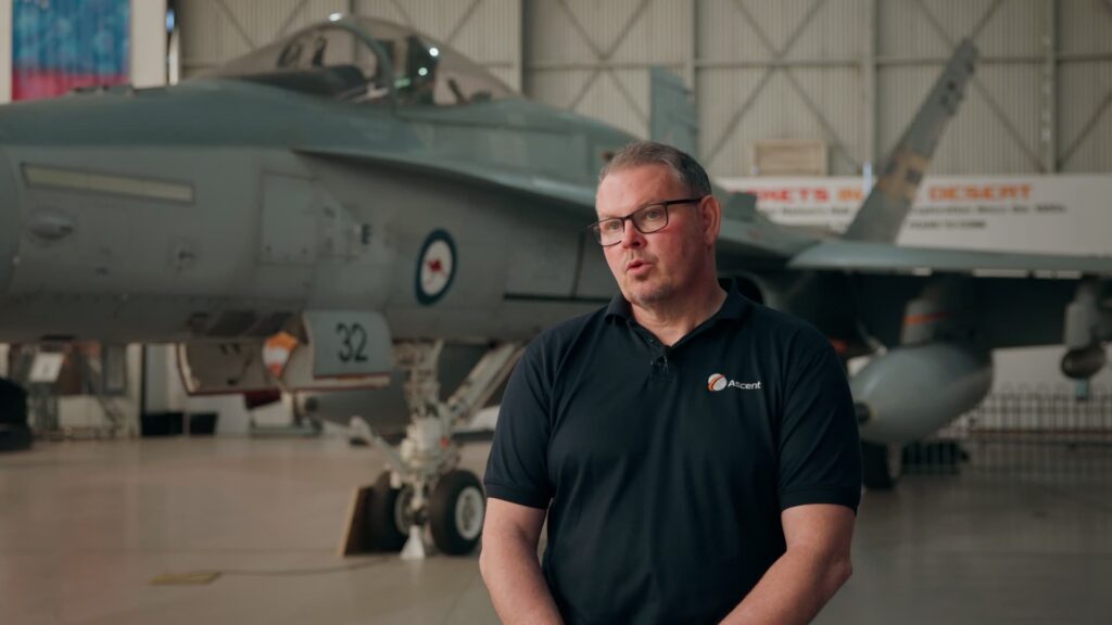 Man in an Ascent-branded polo shirt being interviewed inside an aircraft hangar, with a fighter jet parked in the background.