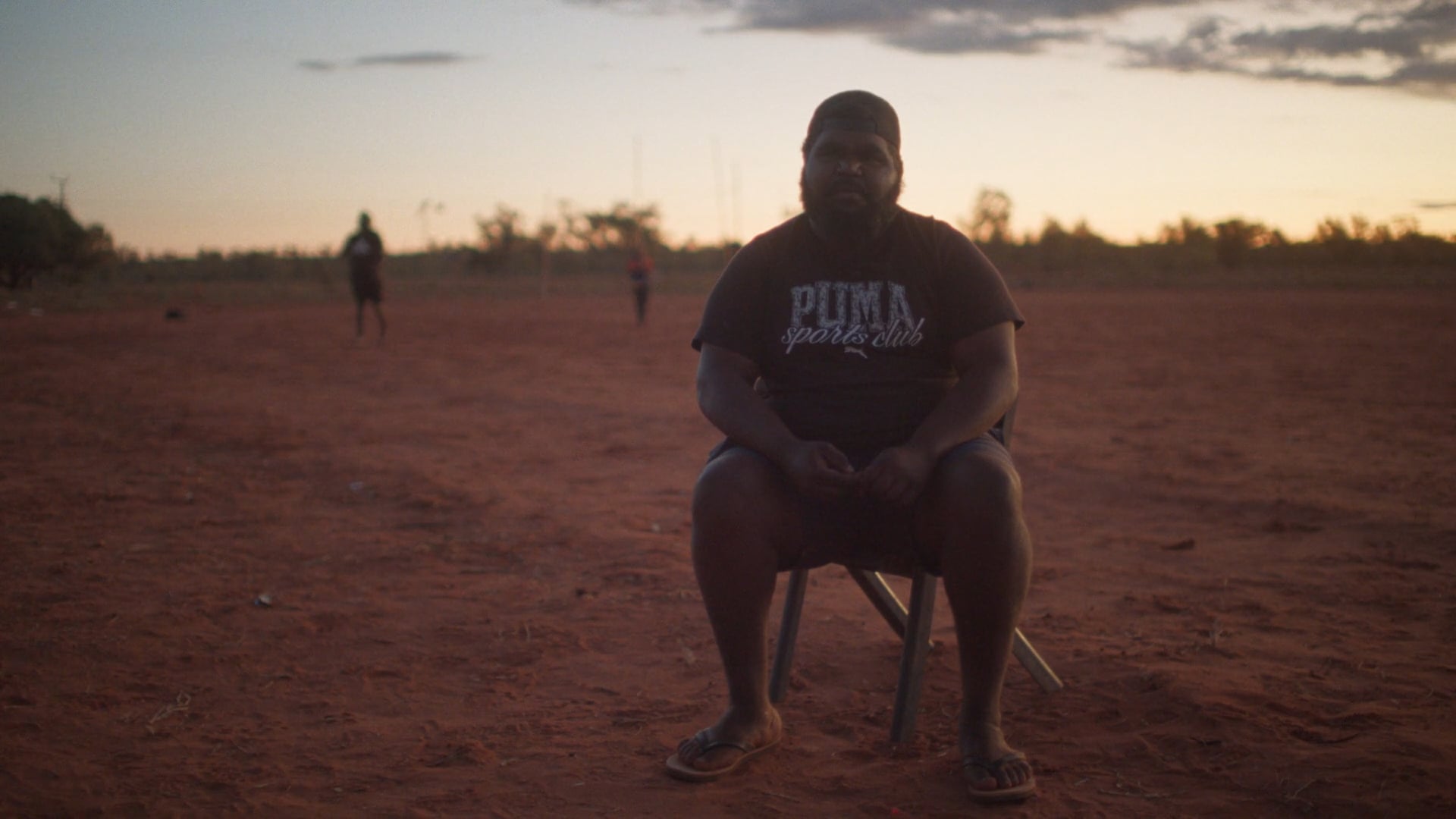 A man sits on a folding chair on a red dirt football field at sunset. The sky glows soft orange and blue, with a few figures in the distance.