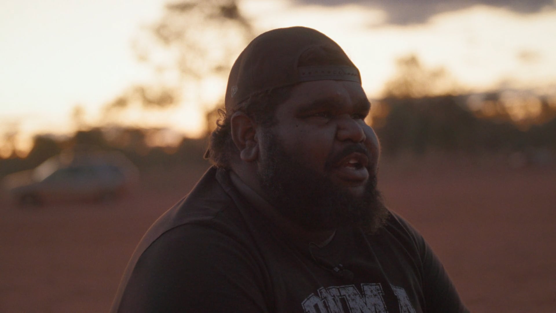 A man sits on a folding chair on a red dirt football field at sunset. The sky glows soft orange and blue, with a few figures in the distance.