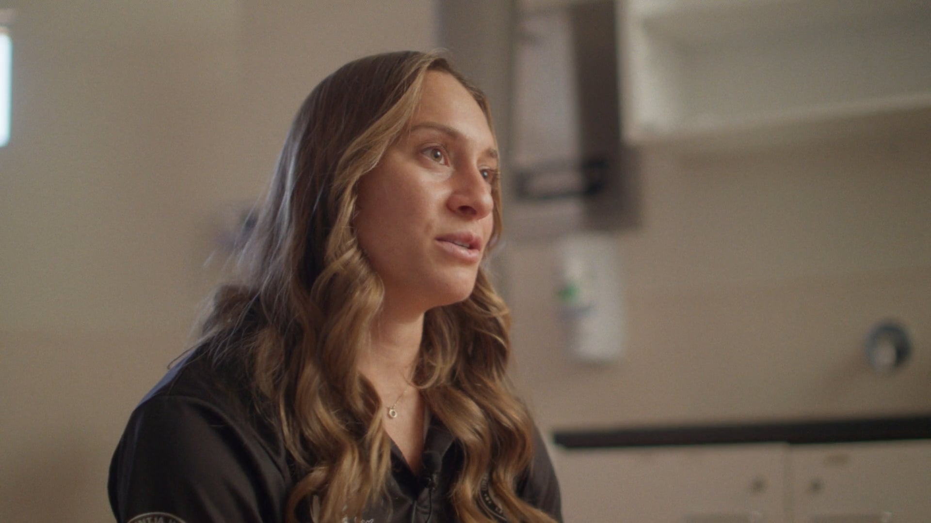 A man sits on a folding chair on a red dirt football field at sunset. The sky glows soft orange and blue, with a few figures in the distance.