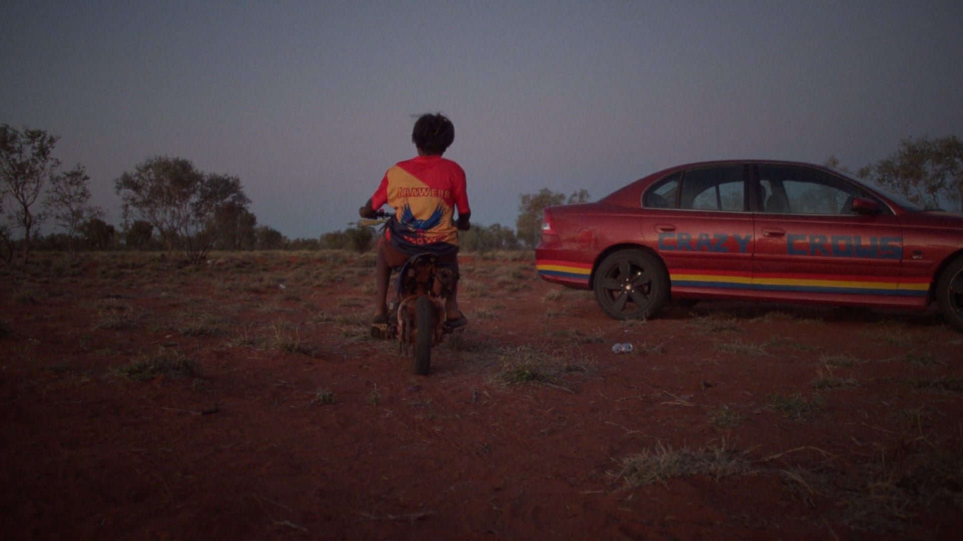 A man sits on a folding chair on a red dirt football field at sunset. The sky glows soft orange and blue, with a few figures in the distance.