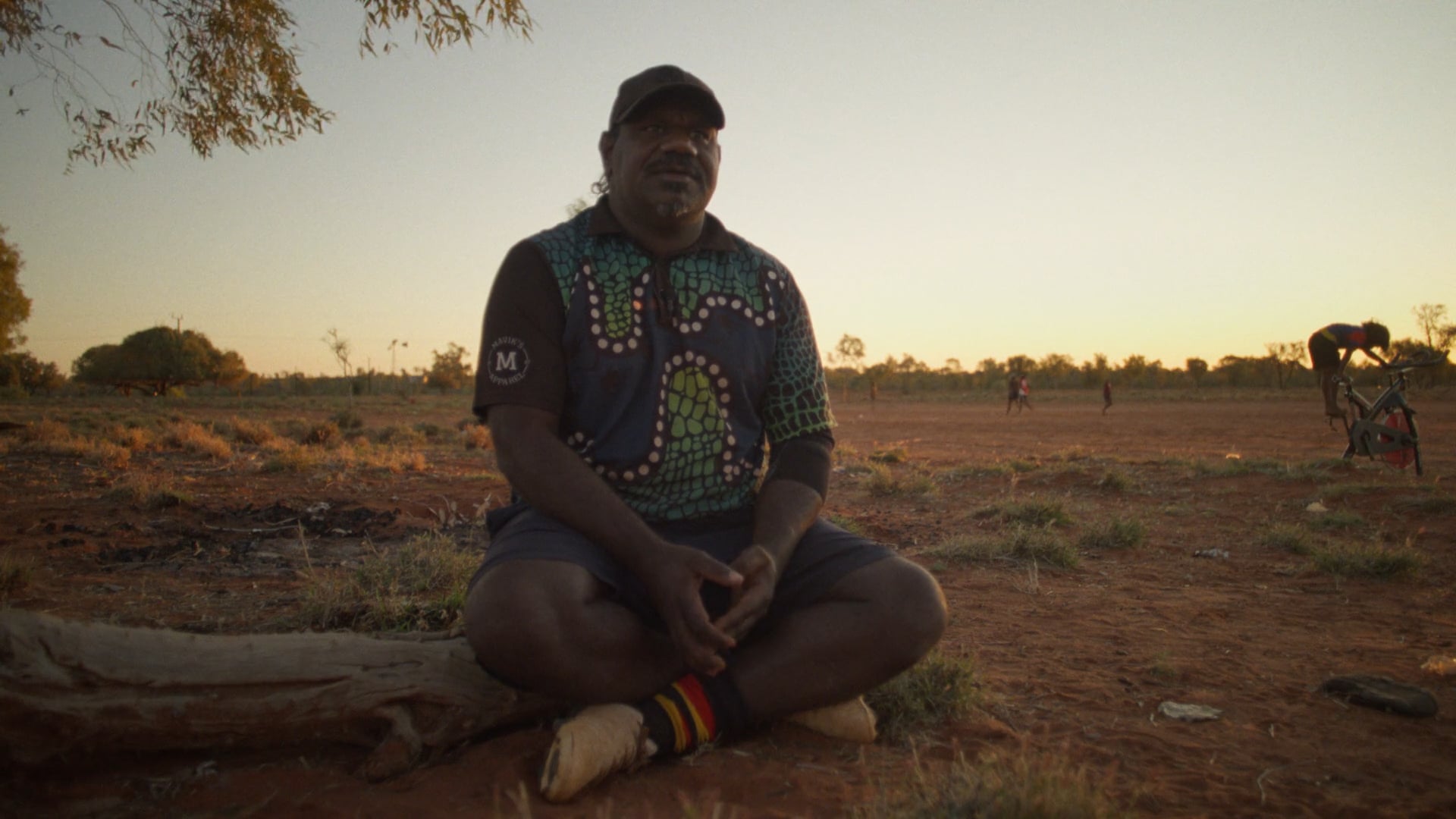 A man sits on a folding chair on a red dirt football field at sunset. The sky glows soft orange and blue, with a few figures in the distance.