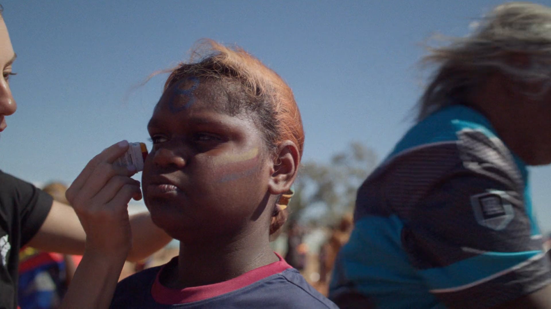 A man sits on a folding chair on a red dirt football field at sunset. The sky glows soft orange and blue, with a few figures in the distance.