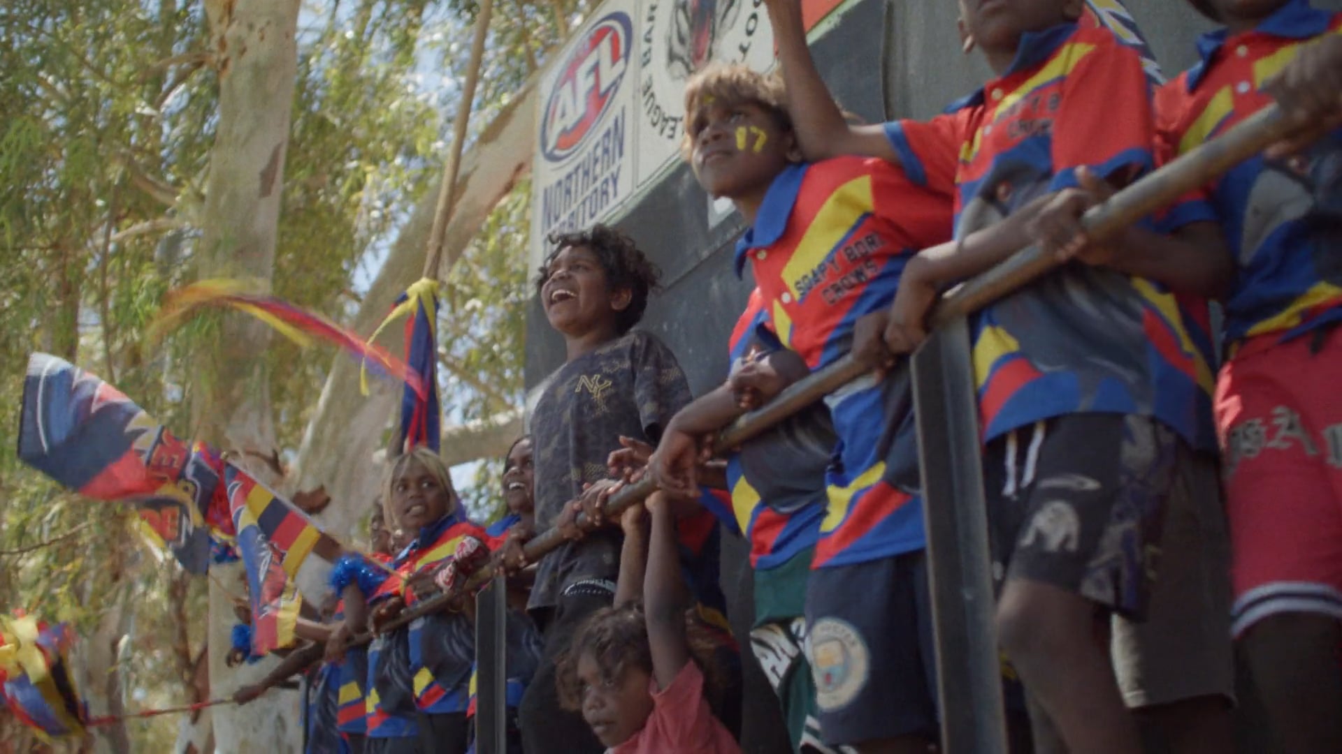 A man sits on a folding chair on a red dirt football field at sunset. The sky glows soft orange and blue, with a few figures in the distance.