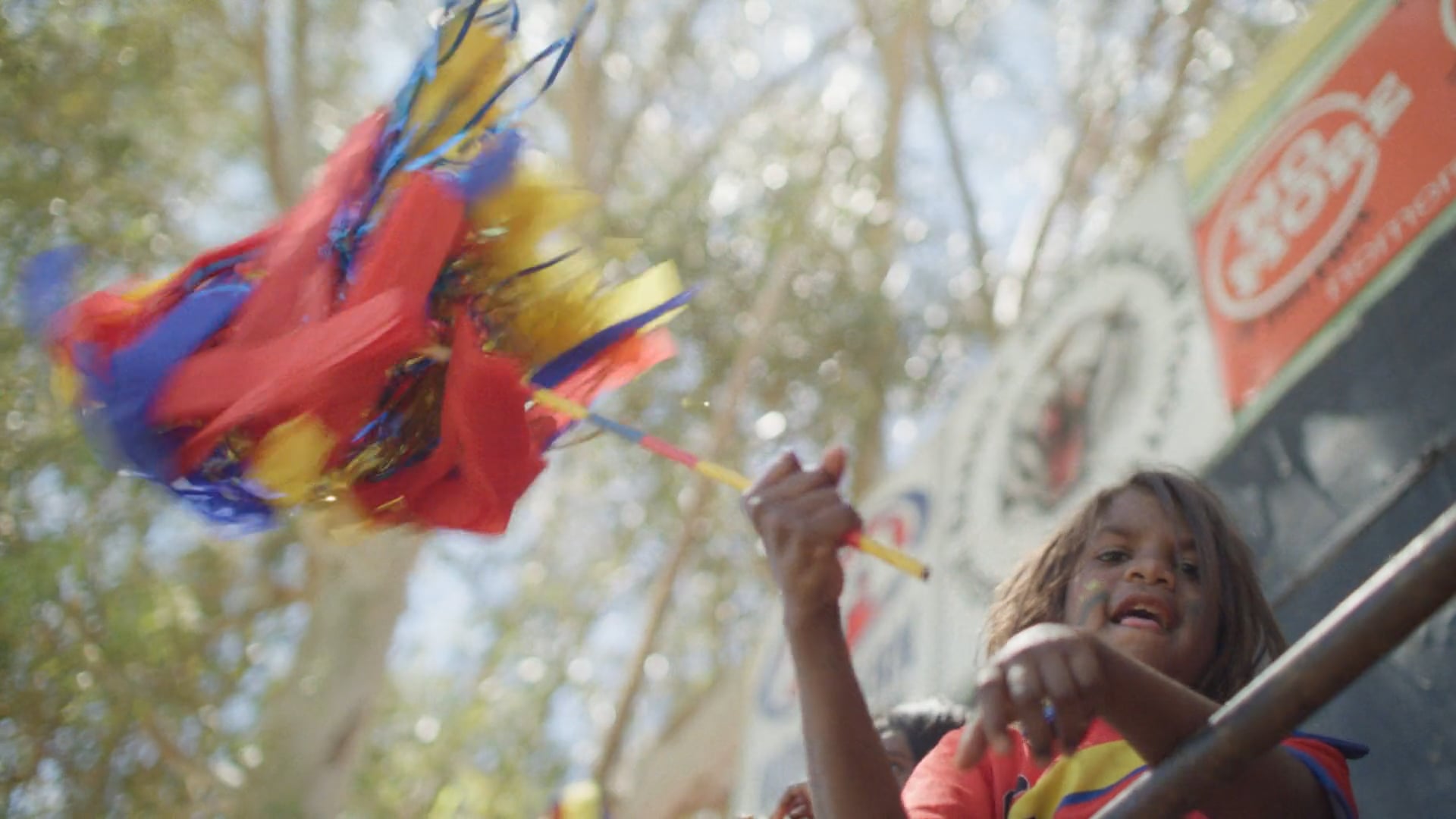 A man sits on a folding chair on a red dirt football field at sunset. The sky glows soft orange and blue, with a few figures in the distance.