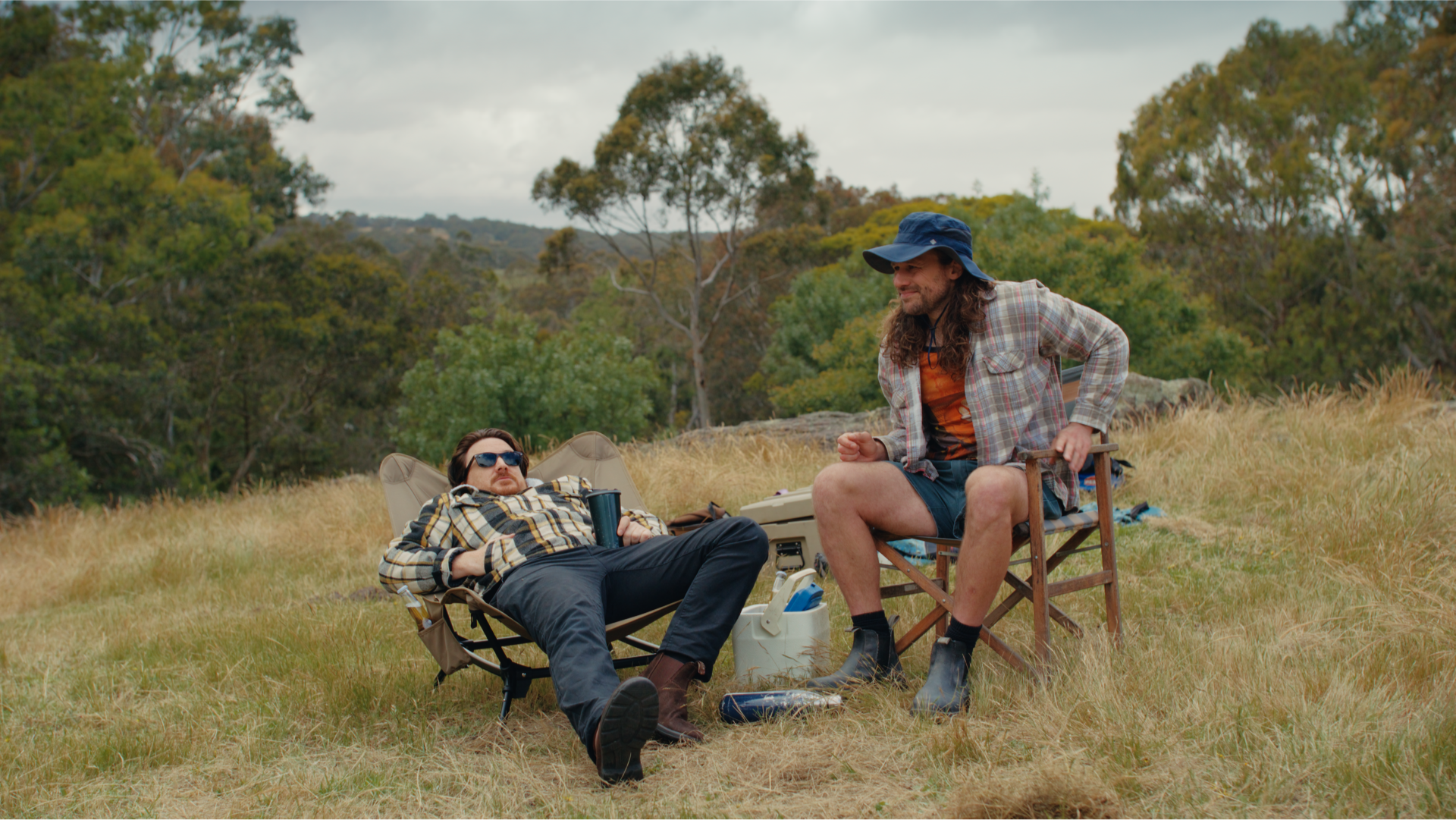 A laid-back camping scene with two men in chairs on dry grass, surrounded by gear and bushland, from Tiegear’s Chair ad.