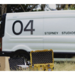 Grainy, lo-fi photo of a white Stepney Studios van marked “04” parked outdoors with camera cases and gear on the grass in the foreground.