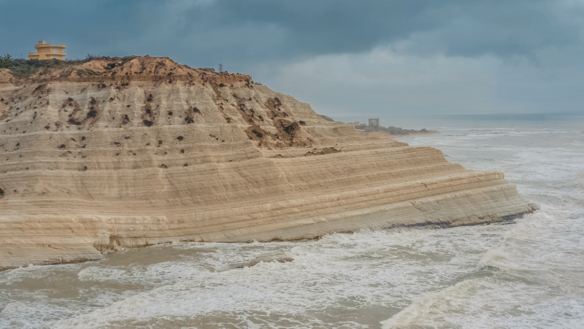 Waves crash against layered sandstone cliffs under a dark, stormy sky, with a small building on the headland.