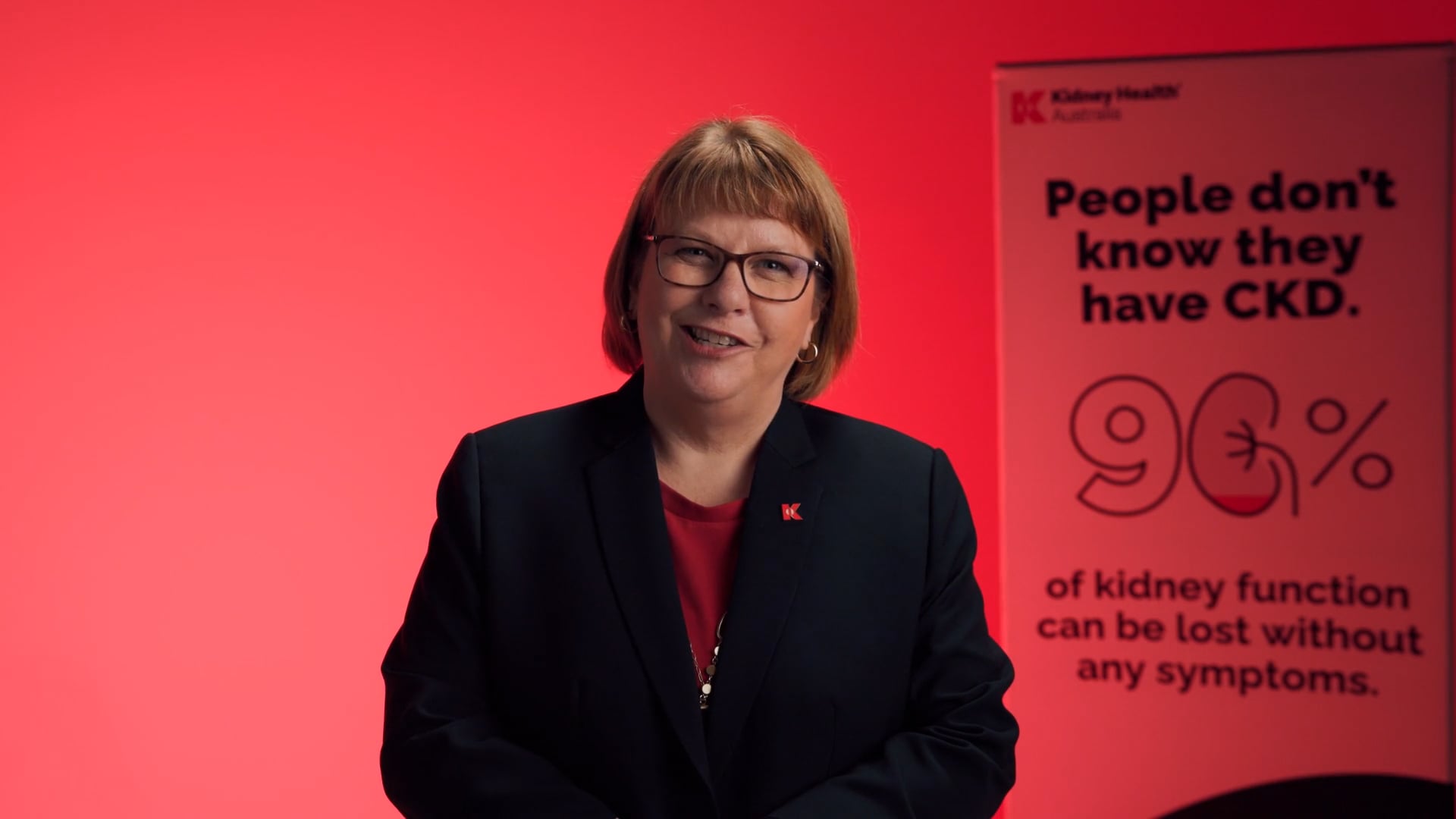 A speaker in a dark blazer sits against a red studio backdrop beside a Kidney Health Australia banner reading “People don’t know they have CKD” and “96% of kidney function can be lost without any symptoms.”
