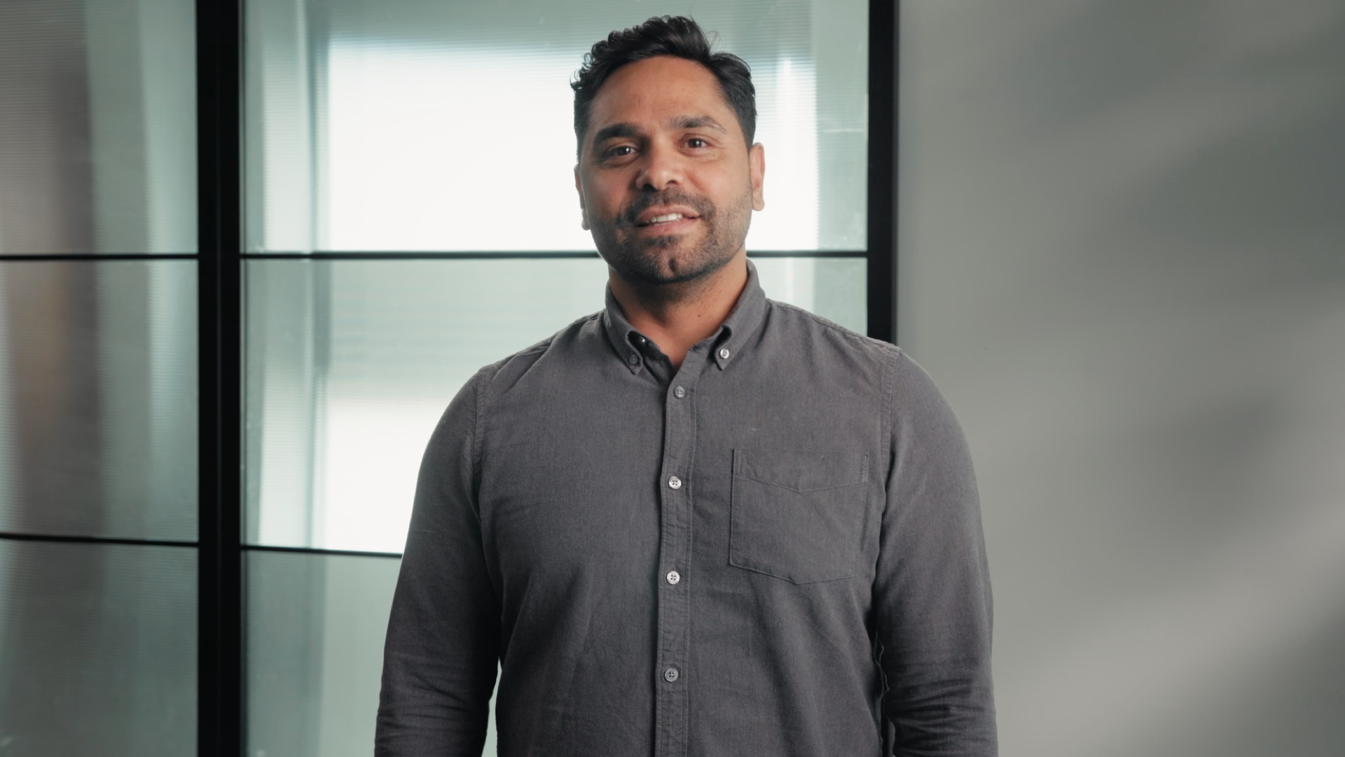 Professional portrait of a man in a grey button-up shirt standing in an office setting, smiling at the camera.