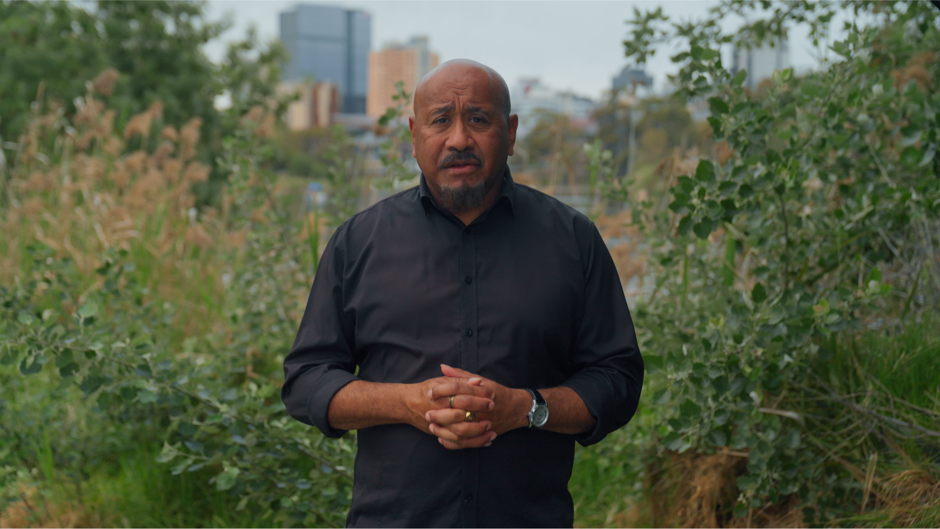 A man in a black shirt speaks to camera outdoors, framed by green foliage with the Adelaide city skyline in the background.