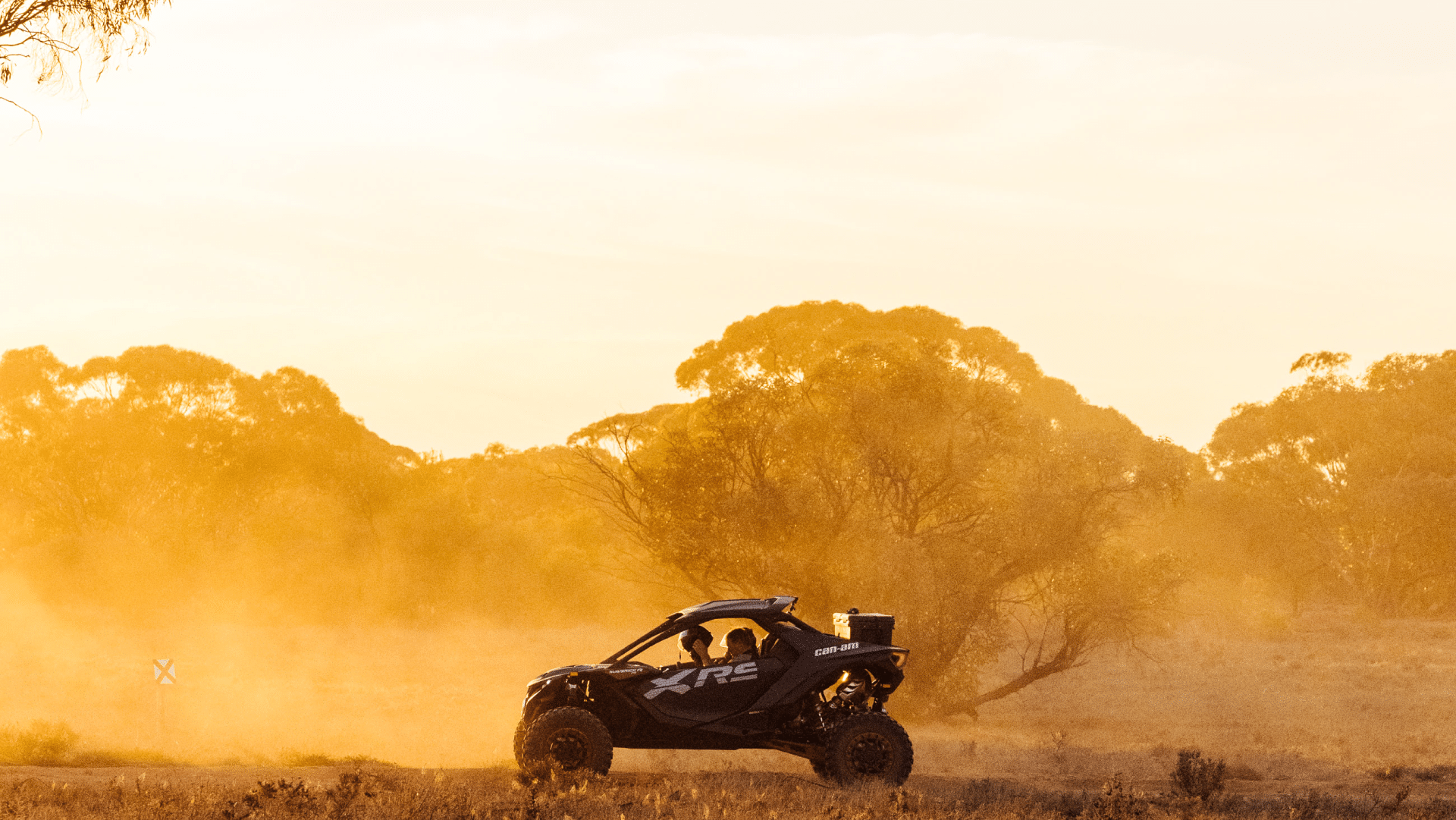 A Can-Am Maverick XRS side-by-side buggy drives through golden outback dust at sunset, with silhouetted eucalyptus trees and a warm orange-lit sky in the background.