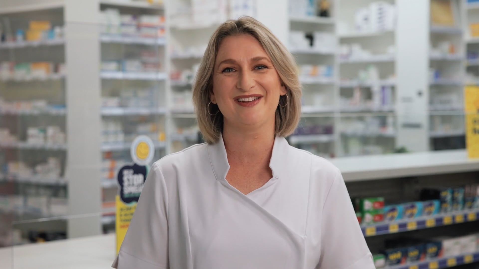A smiling woman in a white pharmacist's uniform stands behind a pharmacy counter, with shelves of health and pharmaceutical products visible in the background.